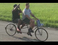 Guy shotguns beer can while he rides backseat on tandem bike