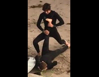 Surfer in wetsuit lifts friends leg up and helps him stretch on the beach