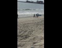 Guy filming behind two girls and guy with surfboard on beach