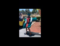 Girl in blue graduation cap and gown dances and trips backwards over curb