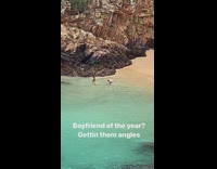 Woman in black bikini poses while man takes picture at the beach