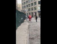 Red monster mascot walks down the side of the road by itself, green tarp fence