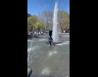 Guy riding bike in water fountain balancing trash can on head