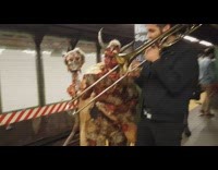 Guy plays the trumpet next to person dressed up as a red demon in subway station