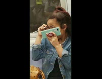 Woman holds spoon to eye and puts mascara on with blue phone as mirror on subway train