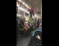 Woman in colorful quilt patch dress and pink purple headpiece stands on seat and hangs on rail on subway train