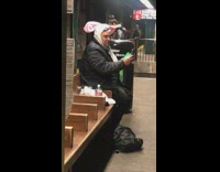 The Jolly Cat plays green tambourine and sings into phone in subway station