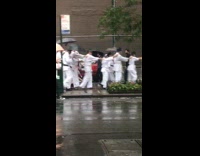 Karate students follow each other in a single file line down the sidewalk in the rain, wearing blindfolds
