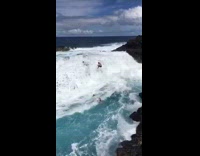 Group of people on top of rocks on beach get pushed into water by waves
