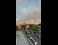 Pride parade going through the streets under a rainbow in the sky 
