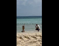 Collab IITW - Woman in bikini kneel while man in gray shirt takes picture from behind at the beach