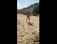 Guy in sunglasses and orange shorts jumps on blue medicine ball in sand, front flips but lands on side