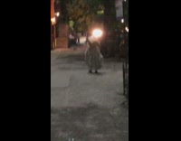 Ghost with white bedsheet blanket and cardboard box on head walks down sidewalk alone at night