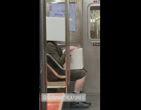 Guy wears big white box over his head and holds another white box on his lap while talking loudly in the corner on a subway train