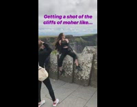 Girl sits on wall and takes picture in front of Cliffs of Moher