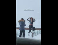 Woman in tight black suit sits on fence and takes picture in snow