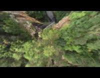 Drone flies down waterfall cliff green trees