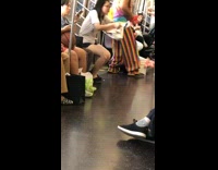 Man in rainbow pants tie dye shirt makes red heart ballon animal for woman on subway train