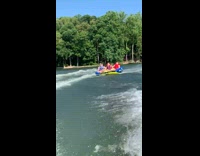 Girl in blue life vest rides on left side of raft, hits a wave and flies off raft into lake water