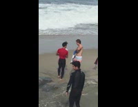 Group of guys carry surfboards down the shore, guy in pastel swimming trunks holds a pink tube