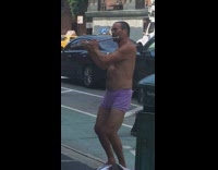 Shirtless man in purple boxers plays the flute and thrusts hips on sidewalk