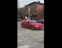 Santa Claus sits on red convertible car, jingles bells and waves at pedestrians
