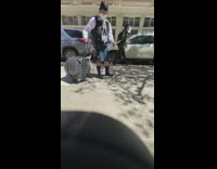 Guy in a kilt walking on the sidewalk dragging a case on wheels 
