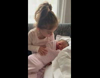 Toddler girl holding her baby sister on couch while baby sleeps