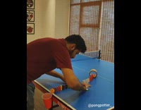 Guy tries to hit ping pong into tape roll and knock down dominoes into red solo cup and fails