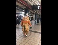 Woman white hair orange floral outfit walks with walking cane around subway station