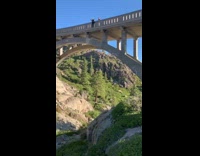 Guy jumps off tall bridge with ropes attached and swings off bridge above rocks