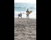 Guy in blue swim trunks takes picture for friends on beach holding yellow surfboard