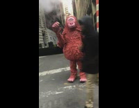 Big red monster mascot waves at the camera and stands on the street next to orange striped steam vent