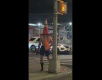 Shirtless guy dances with street pole with cone on his head 