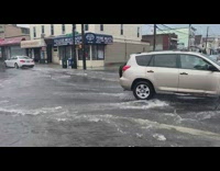 Cars pass by flooded street intersection overflowing storm drain