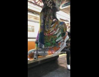 Guy in multicolor quilt dress stands on top of subway seats and holds onto rail