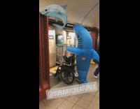 Inflatable blue shark pushes woman in cage wheelchair in subway station